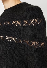 Maglione nero lavorato a maglia con filato testurizzato e dettagli in pizzo lungo le spalle, che aggiungono particolari intricati e contrasto.