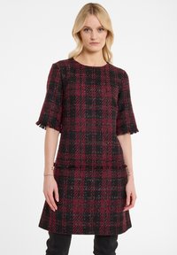 Robe à carreaux rouge et noir en tissu texturé. Présente des manches courtes à franges et une silhouette ajustée avec un ourlet évasé.