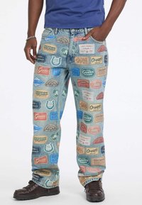 Mężczyzna ubrany w luźne dżinsy z naszywkami kolorowego logo marki Guess, w połączeniu z ciemnobrązowymi butami i niebieską koszulą.