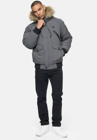 Graue Pufferjacke mit einem mit Futter versehenen Kapuze, Reißverschlusstaschen, gerippten Bündchen und einem bestickten Logo auf der Brust. Getragen mit schwarzen Jeans und weißen Sneakers.