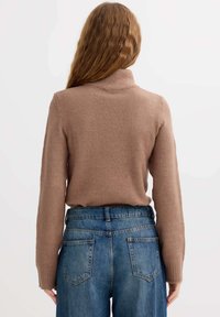 Jersey de punto marrón con cuello alto y mangas largas. Se lleva sobre unos vaqueros de mezclilla azul claro de cintura alta con un diseño clásico de cinco bolsillos.