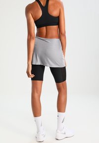 Sujetador deportivo negro, falda envolvente gris sobre pantalones cortos de ciclismo negros, zapatos deportivos blancos y calcetines blancos con un patrón. Tela lisa, diseño ajustado.