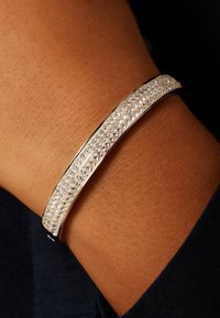 Zilveren armband met een strak ontwerp, voorzien van een rij heldere kristallen die in het midden zijn ingelegd, gedragen aan een pols.