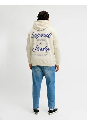 Person trägt einen cremefarbenen Hoodie mit blauem Text "Originals Studio" auf dem Rücken, blaue Jeans und schwarz-weiße Sneakers, steht mit dem Rücken zur Kamera.