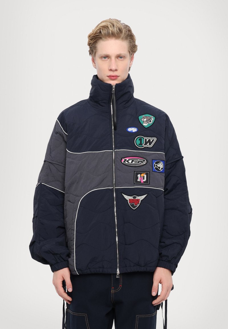 Navy and gray padded jacket with high collar, front zipper, and multiple colorful patches; features a quilted texture and distinct stitching.