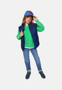Blauwe puffer vest over een groene sweatshirt, in combinatie met casual jeans en donkere sneakers. Voorzien van een blauwe pet met rode logo-accent.
