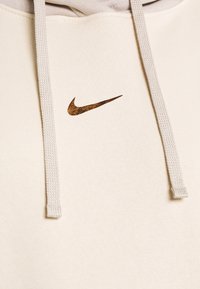 Ljusbeige hoodie gjord av mjukt tyg, med en texturerad brun Nike-logotyp och grå snoddar. Enkla linjer med ingen mönstring.