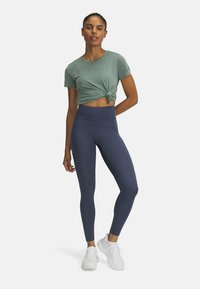 T-shirt annodato verde chiaro abbinato a leggings blu scuro a vita alta. Tessuto liscio e elastico con sneaker bianche. Design semplice e aderente.