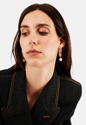Boucles d'oreilles en or avec un détail en perle. Le modèle porte un blazer en denim foncé avec des coutures contrastées. Gros plan sur le visage et les accessoires.