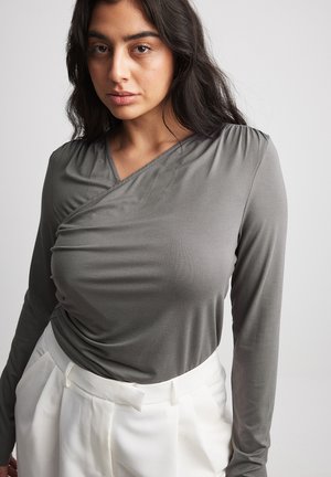 Camiseta de manga larga gris con un diseño envolvente y tejido texturizado. Combinada con pantalones blancos de cintura alta, que presentan un detalle de cinturón.