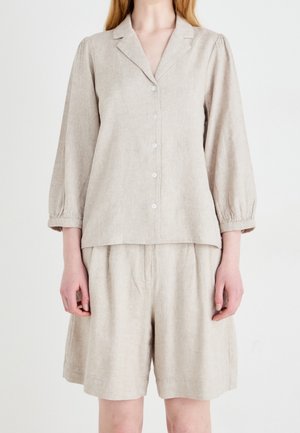 Beżowa koszula na guziki z długim rękawem i kołnierzykiem, zestawiona z dopasowanymi luźnymi szortami na osobie na białym tle.