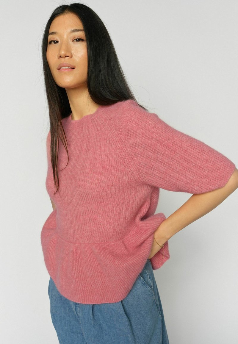 Femme aux longs cheveux noirs portant un pull rose duveteux et un jean bleu, posant avec une main sur la hanche devant un fond uni.