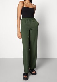 Zwarte spaghetti-top gecombineerd met losse donkergroene broek met elastische tailleband en zijzakken. Zwarte platformsandalen.