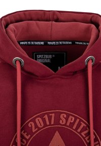 Burgunderfarbener Kapuzenpullover mit rundem Logo, das "SPITZBUB" und das Jahr "2017" zeigt. Zugbänder und Nähte in einer helleren Nuance, aus weichem Textilmaterial.