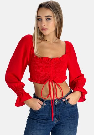 Rote, verkürzte Bluse mit gerüschtem Muster, quadratischem Ausschnitt und langen Puffärmeln. Hat eine Bindeschleife vorne und fransige Kanten.