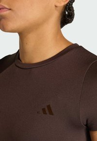 Καφέ αθλητική μπλούζα με ριμπ υφή, στρογγυλεμένο ντεκολτέ και διακριτική σκούρα καφέ λεπτομέρεια λογότυπου στο στήθος. Κοντινή λήψη, πλάγια όψη.