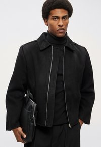 Crna vunena jakna s patentnim zatvaračem, velikim ovratnikom i veznim detaljima, uparena s crnim rolkom i držeći crnu kožnu torbu.