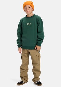 Zelený mikina s logom, kombinovaná s khaki cargo nohavicami s vreckami. Doplnené: čierno-biele tenisky, jasne oranžová čiapka.