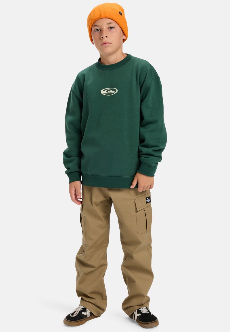 Zelený mikina s logom, kombinovaná s khaki cargo nohavicami s vreckami. Doplnené: čierno-biele tenisky, jasne oranžová čiapka.