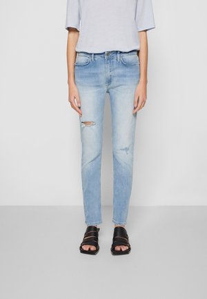 Person iført lyseblå slidte jeans, lysegrå kortærmet top og sorte åbne tå-sandaler, der står mod en ensfarvet baggrund.