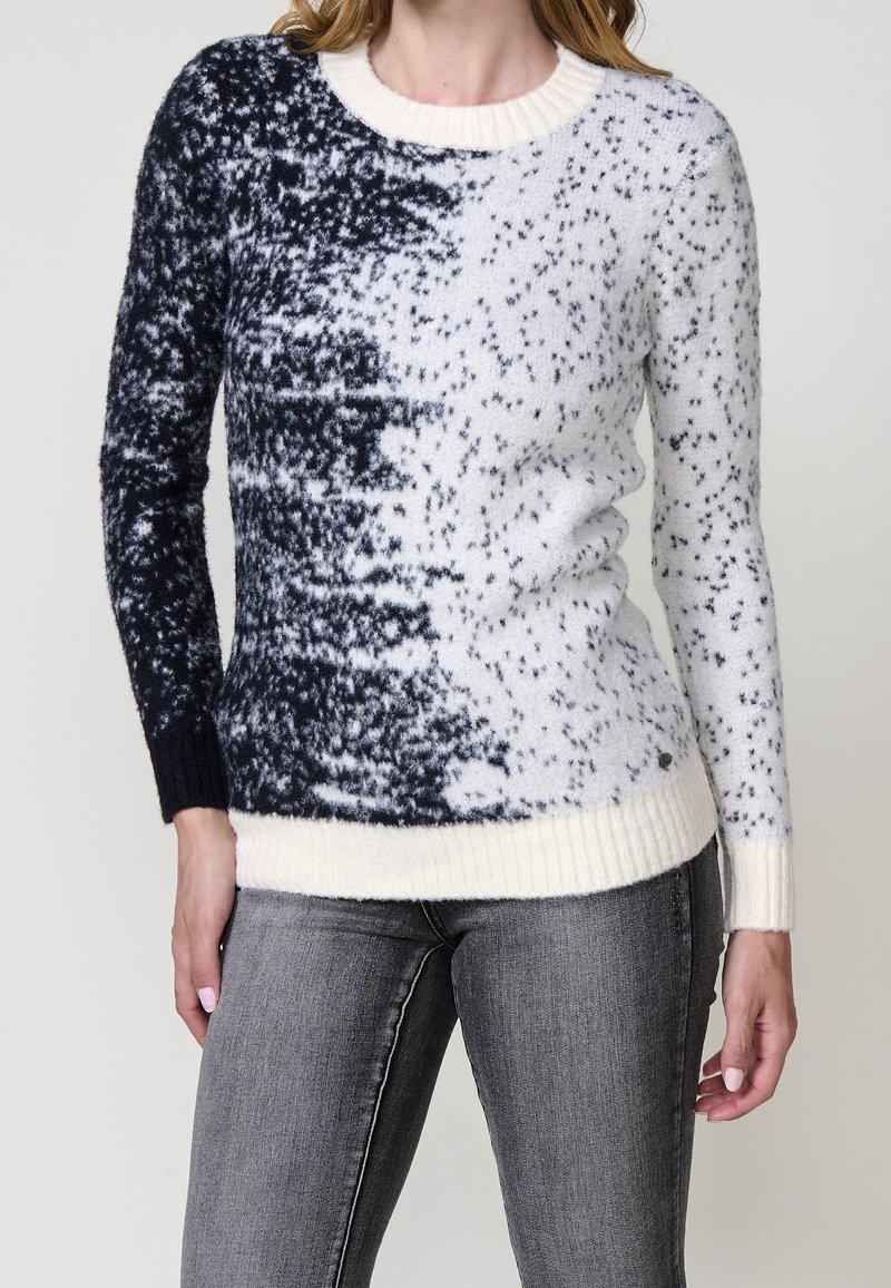 Femme portant un pull en maille à manches longues avec un motif moucheté noir et blanc et des poignets et ourlet côtelés, associé à un jean gris.