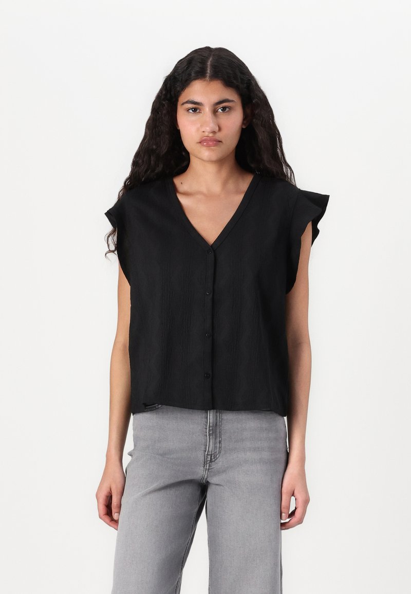 Svart kortermet bluse med V-hals og knappestolpe, med et teksturert mønster og en avslappet passform. Kombinert med grå jeans.
