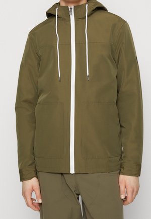 Homme portant une veste à capuche vert olive avec zip blanc et cordons, associée à un pantalon vert olive assorti.