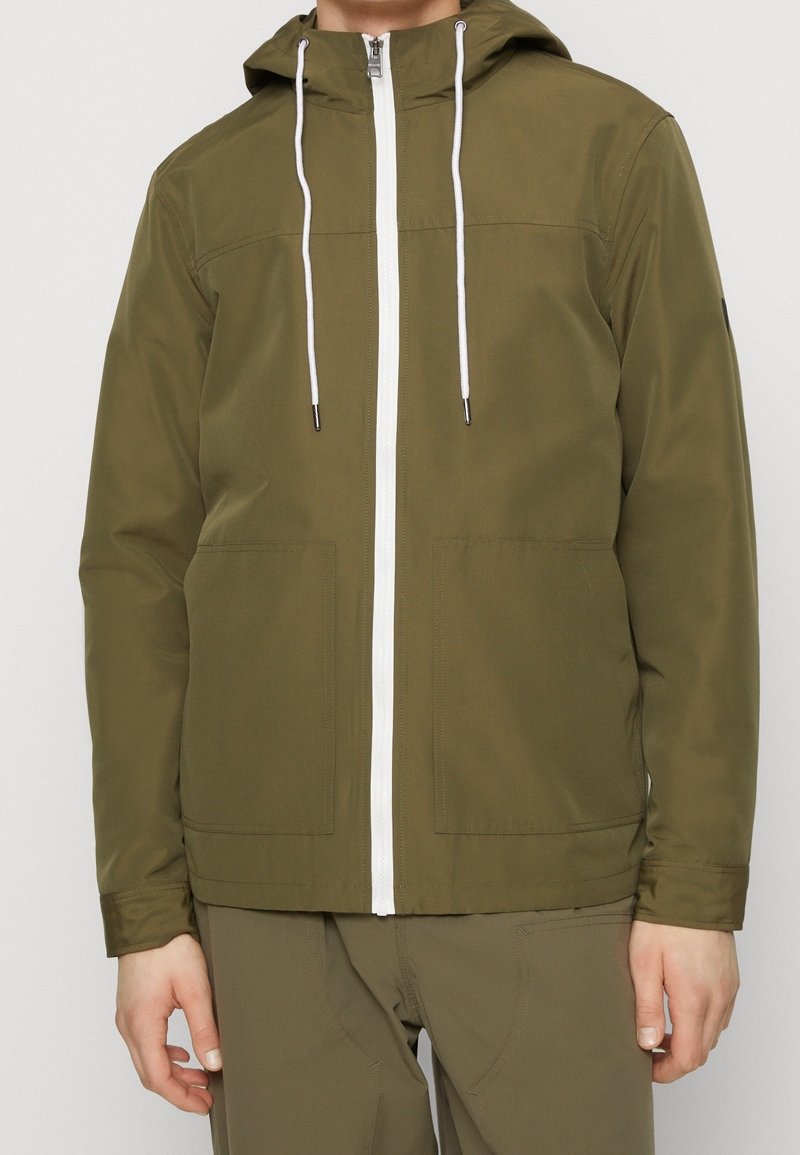 Homme portant une veste à capuche vert olive avec zip blanc et cordons, associée à un pantalon vert olive assorti.