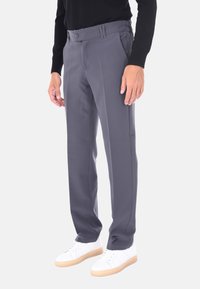 Uomo in pantaloni dritti grigi con pince frontali e sneaker bianche basse, in piedi di fronte a uno sfondo neutro.