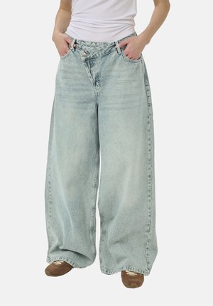 Person wearing light blue wide-leg jeans with an asymmetrical buttoned waistband and brown sneakers, hands in pockets.