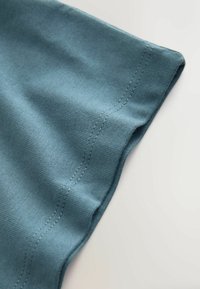 Puuvillaisen t-paidan hiha pehmeässä teal-värissä, jossa on sileä pinta ja siististi ommellut reunat. Kangas näyttää kevyeltä ja joustavalta.