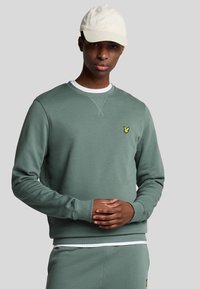 Felpa verde con polsini e colletto a coste, con un piccolo logo giallo sul petto. Il modello indossa un cappellino chiaro.
