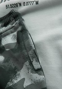 Szara bluza z czarnymi współrzędnymi, zestawiona z ciemnozielonymi spodniami dresowymi w kamuflażu, które mają teksturowaną powierzchnię i detal bocznej kieszeni.