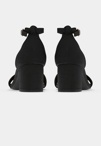 Mustat korkokengät, joissa on suljettu takaosa, nilkkaremmi ja avoin varvasdesign. Varusteena leveä lohkokorko ja sileä tekstuuri.