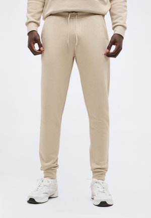 Homme portant un pantalon de survêtement beige et un sweat-shirt avec des baskets blanches, debout devant un fond clair uni.