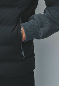 Asmuo, vilkintis tamsiai mėlyną pūkinę striukę su priekiniais užtrauktukais, ranką įsidėjęs į šoninį kišenę, o matoma pilka mezgimo rankovė.