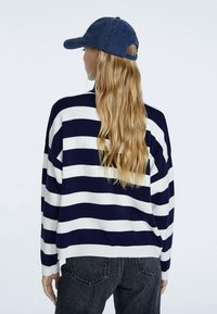 Navy en witte gestreepte trui met een ontspannen pasvorm, geribbelde manchetten en een detail op de rug. Gepaard met een marineblauwe baseballcap en donkere jeans.
