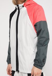 Veste légère composée d'un mélange de panneaux blancs, corail et gris foncé, avec une fermeture éclair à l'avant, une capuche ajustable et des poignets élastiques.