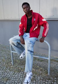 Giacca da track rossa con strisce bianche, t-shirt nera, jeans larghi azzurri chiari e sneakers bianco e nere. Seduto su una ringhiera metallica.