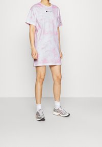 Różowa i lawendowa sukienka t-shirt w stylu tie-dye z krótkimi rękawami, z małym czarnym logo na klatce piersiowej, noszona z srebrnymi butami sportowymi.