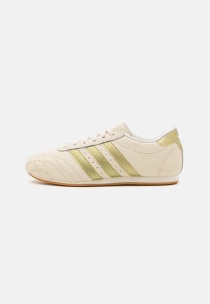 Kremfargede lær sneakers med tre metalliske gullstriper, teksturert tåboks og en tan gummisåle. Har perforerte detaljer på sidene.