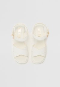 Sandales blanches avec des brides croisées et un bout carré, présentant un accent de boucle en or sur la bride de la cheville. Texture mate et lisse.
