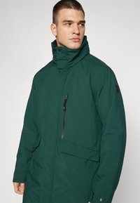 Grønn, isolert jakke med høy krage, med en glidelåslomme på brystet og knappefeste. Glatt tekstur med lett design.