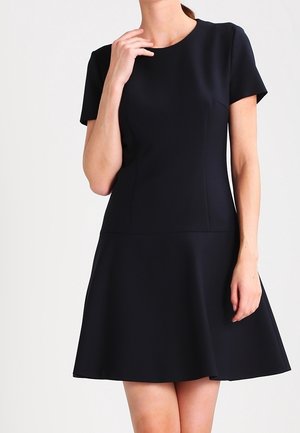 Vrouw draagt een zwarte jurk tot op de knie met korte mouwen, een nauwsluitend lijfje en een uitlopende rok, tegen een effen achtergrond.