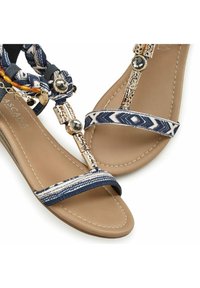 Blaue Sandalen mit Muster, die eine flache Sohle, dekorative Riemen, goldfarbene Akzente und ein geflochtenes Design mit Perlen aufweisen.