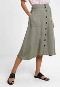 Khaki midi-hame flare-mallisella, nappi edessä, kaksi taskua ja sileä pinta. Yhdistetty mustiin tasapohjaisiin sandaaleihin.