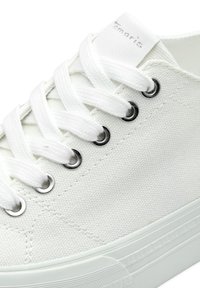Białe trampki z płótna z metalowymi oczkami, płaskimi białymi sznurówkami i fakturowaną powierzchnią tkaniny. Posiadają solidną białą gumową podeszwę.