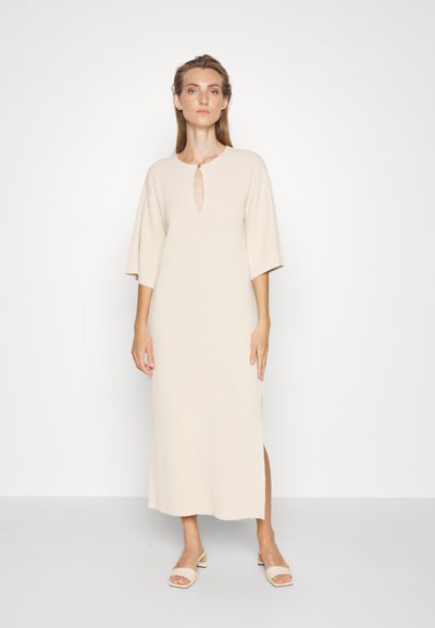 Φόρεμα σε μπεζ απόχρωση μέχρι το γόνατο, μαλακή υφή, λαιμόκοψη σε σχήμα κλειδώματος, χαλαρές κοντές μανίκες, πλευρικό άνοιγμα, φορεμένο με ανοιχτόχρωμα παπούτσια με ανοιχτές μύτες.