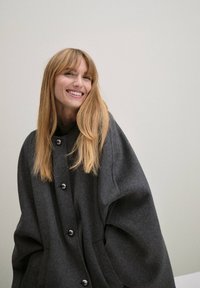 Manteau en laine gris surdimensionné avec des boutons ronds, en tissu légèrement texturé, et un design fluide. Caractéristiques d'un col haut et de manches drapées.