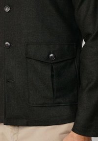 Donker visgraatjack met een knoopsluiting aan de voorkant, een borstzak met klep en een gestructureerde stof. De knopen zijn zwart.